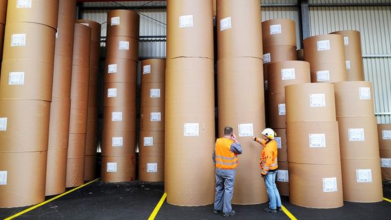Due lavoratori con giubbotti di sicurezza arancioni si trovano davanti a rotoli di carta in una cartiera. Due lavoratori con giubbotti di sicurezza arancioni si trovano davanti a rotoli di carta in una cartiera. © picture alliance / dpa Foto: Jonas Gutler