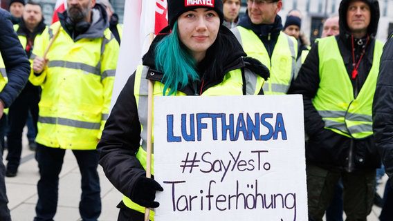 Un dipendente tiene un cartello con la scritta Lufthansa #SayYesTo per un aumento della tariffa durante un raduno del dipartimento Lufthansa Technik di fronte alla sala partenze del Terminal 1 dell'aeroporto BER.  © Carsten Kohl/DPA 